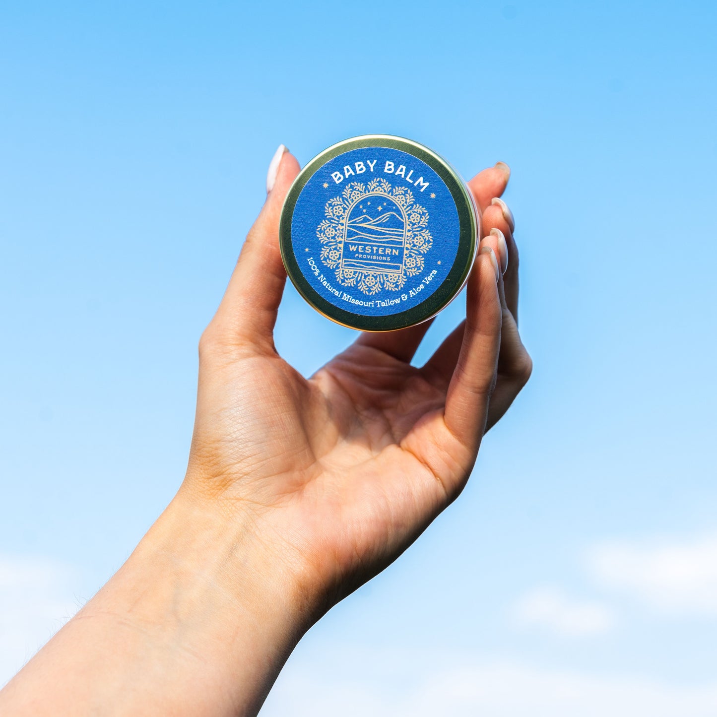 A hand holding Western Provisions tallow baby balm against a blue sky