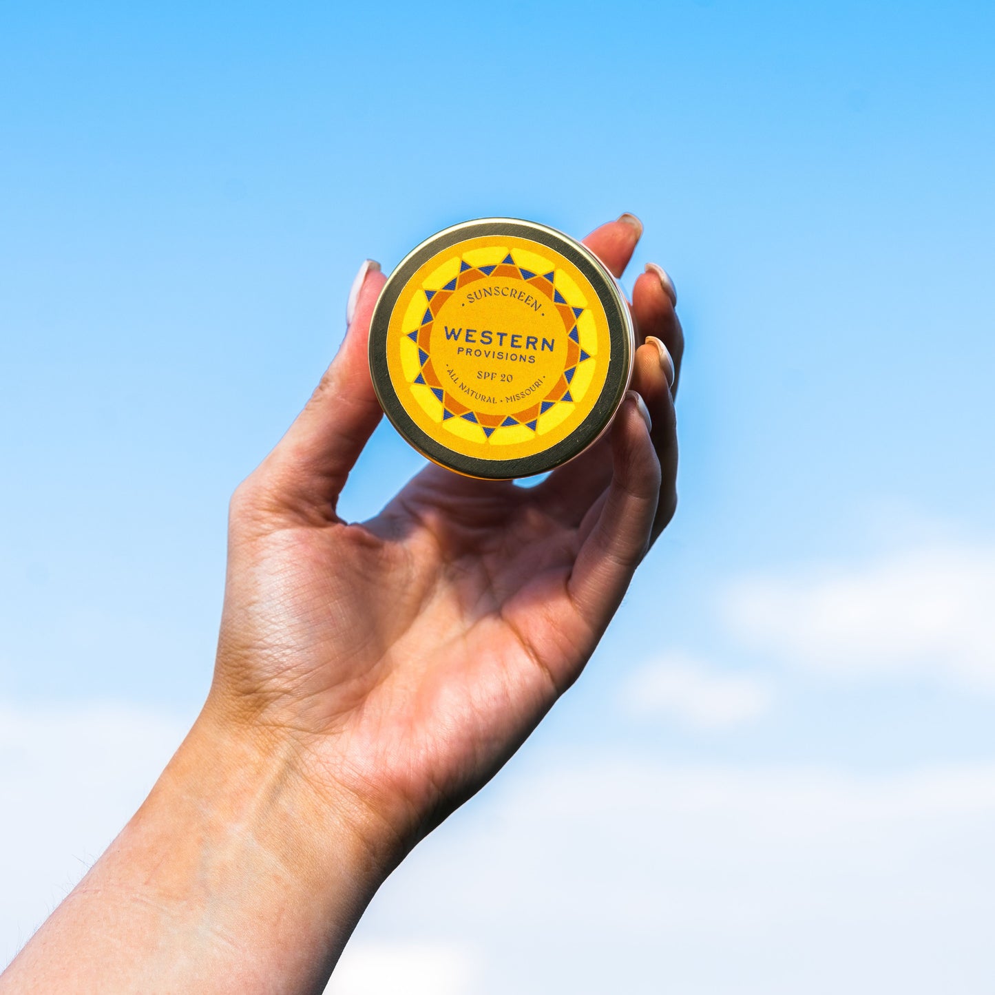 A hand holding Western Provisions tallow sunscreen against a blue sky